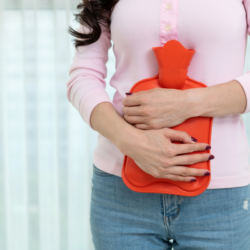 Une femme tenant une bouillotte en caoutchouc rouge contre son abdomen pour soulager la douleur. Image illustrant l'usage thérapeutique de la bouillotte en naturopathie, notamment pour apaiser les douleurs menstruelles et favoriser la détente.