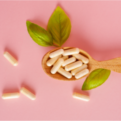 Une cuillère en bois contenant des gélules blanches sur un fond uni rose avec une feuille verte de basilic à côté, symbolisant les compléments alimentaires bio et naturels.