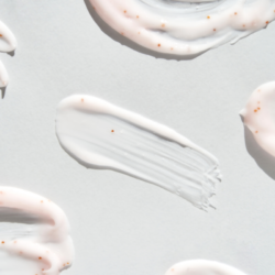 Image en gros plan de différentes traînées de crème et d'exfoliant avec de petites particules marron, sur un fond blanc éclatant. Ces textures crémeuses et ces grains évoquent les composants naturels souvent mentionnés dans les listes d'ingrédients INCI des produits de beauté biologiques.