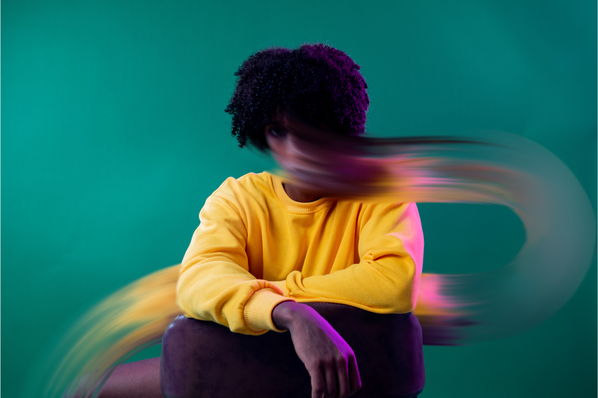 Portrait d'une personne assise, vêtue d'un pull jaune vif, avec une traînée floue de couleurs qui traverse l'image, évoquant un sentiment de mouvement ou de tourbillon d'anxiété, sur un fond vert uni.