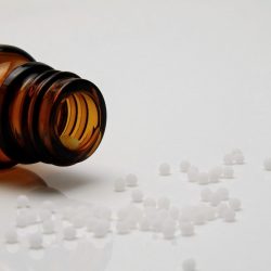 L'image représente un flacon ambré renversé. Des granules d'homéopathie déversées sur une surface blanche.