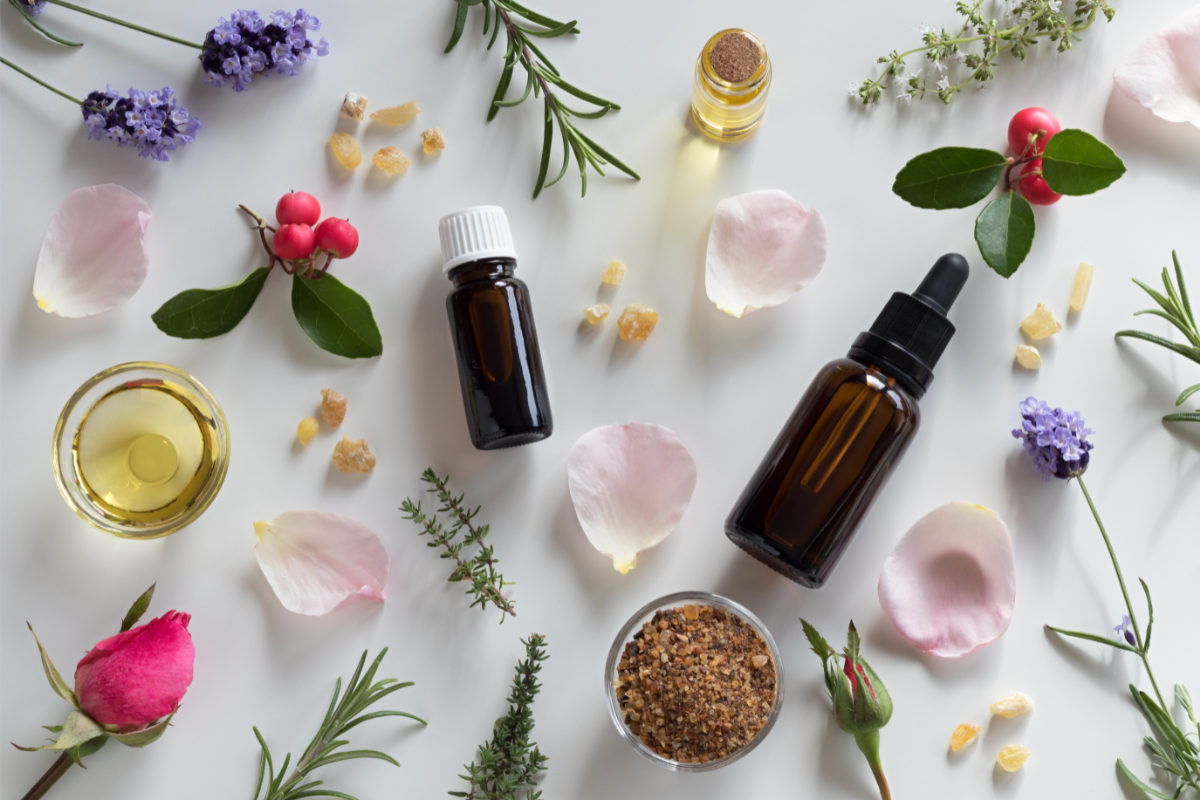 Une composition harmonieuse de produits d'aromathérapie et d'éléments naturels sur un fond blanc. Plusieurs flacons en verre brun, dont certains équipés de pipettes, contenant des huiles essentielles, sont dispersés parmi des pétales de rose pâle et de rose vif, des branches de lavande, des feuilles de romarin, des baies rouges, des pépites de résine d'ambre et un petit bol de cristaux de sel brun. L'image dégage une ambiance de sérénité et d'équilibre naturel, évoquant les principes et la douceur de l'aromathérapie pratiquée en naturopathie.