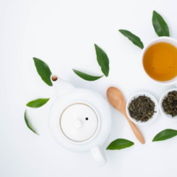 Cette photographie aérienne présente un ensemble d'objets associés à la consommation du thé vert. Au centre, une théière blanche en céramique avec un couvercle en bois est mise en évidence sur un fond blanc épuré. À sa droite, une tasse blanche contient du thé vert infusé, dont la couleur ambre légère contraste avec la blancheur de la porcelaine. Trois petits bols également en porcelaine, accompagnés de cuillères en bois, contiennent des feuilles de thé vert séchées, variant légèrement en texture et en teinte. Éparpillées avec esthétique, des feuilles fraîches de thé vert ajoutent une touche de verdure vive, complétant la composition par leur couleur et leur fraîcheur.