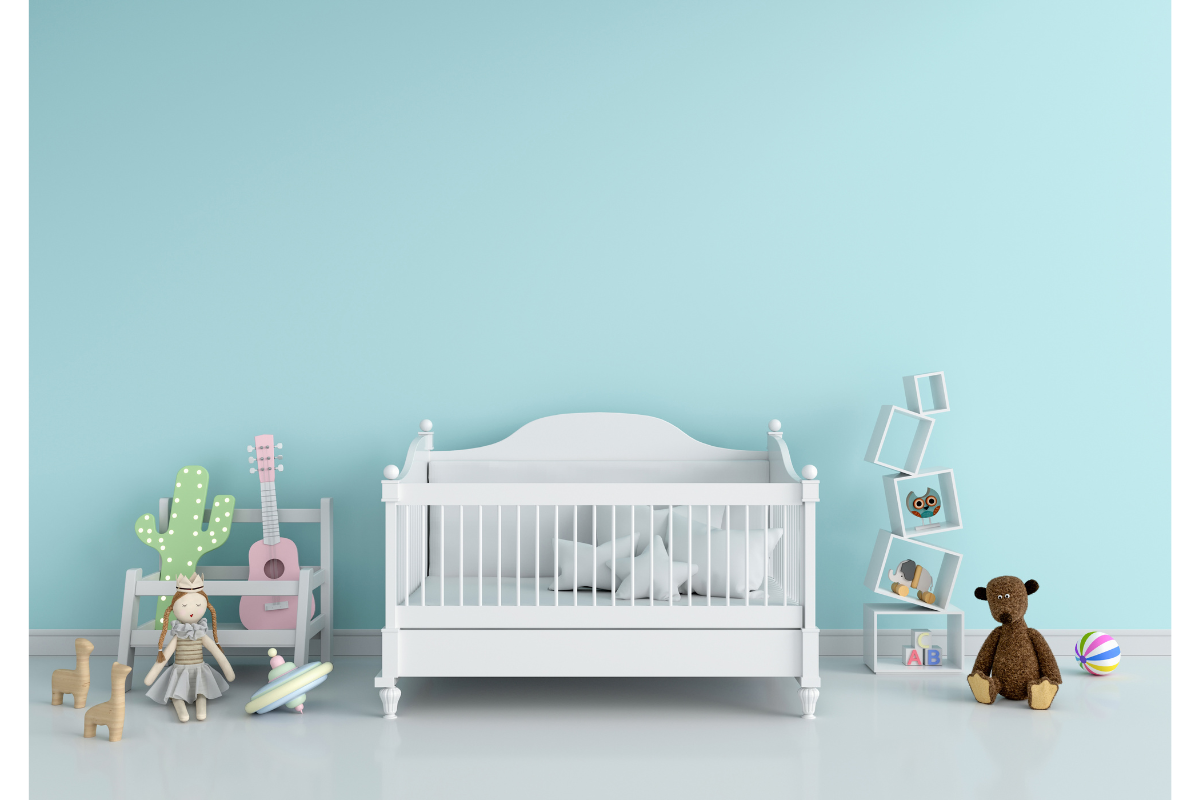 Image d'une chambre d'enfant paisible et minimaliste avec un mur d'un bleu pastel doux. Au centre, un berceau blanc classique avec des draps blancs évoque une atmosphère de calme et de sécurité pour le nourrisson. Sur la gauche, une étagère en forme de cactus et une guitare rose miniature suggèrent une ambiance ludique et créative. À droite, des blocs de construction avec des lettres alphabétiques et un cadre contenant une illustration de hibou renforcent un environnement éducatif. Un ours en peluche brun et un ballon multicolore sont posés sur le sol, ajoutant une touche de confort et d'invitation au jeu. L'ensemble crée une atmosphère sereine qui peut contribuer à l'apaisement des coliques du nourrisson grâce à un environnement stable et réconfortant.