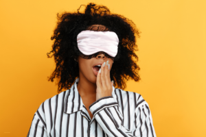 Femme en pyjama avec un masque de sommeil rose bâillant devant un fond jaune, symbolisant les troubles du sommeil et les solutions naturelles comme les Fleurs de Bach.