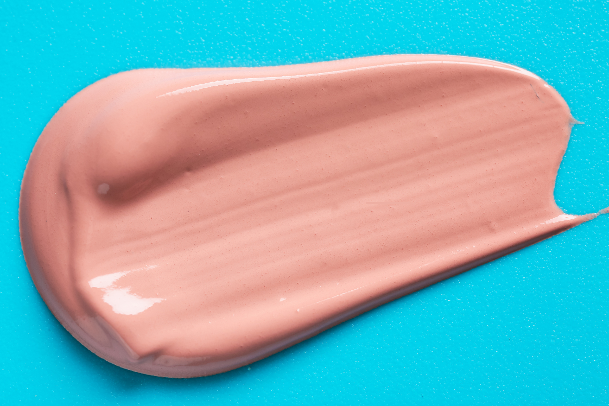 Échantillon de BB crème de couleur beige rosé sur fond bleu, illustrant la texture fluide et couvrante d’un soin hybride alliant hydratation et correction du teint.