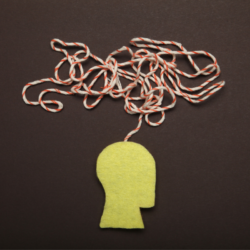 Une illustration symbolique montrant une silhouette de tête humaine en feutre jaune avec un fil rouge et blanc enchevêtré émergeant de son sommet, sur un fond marron foncé. Cela représente métaphoriquement la complexité des pensées et des émotions humaines durant la période de rentrée, et comment les compléments alimentaires, comme la créatine, peuvent être envisagés pour soutenir la santé mentale et cognitive des adultes face à ces défis.