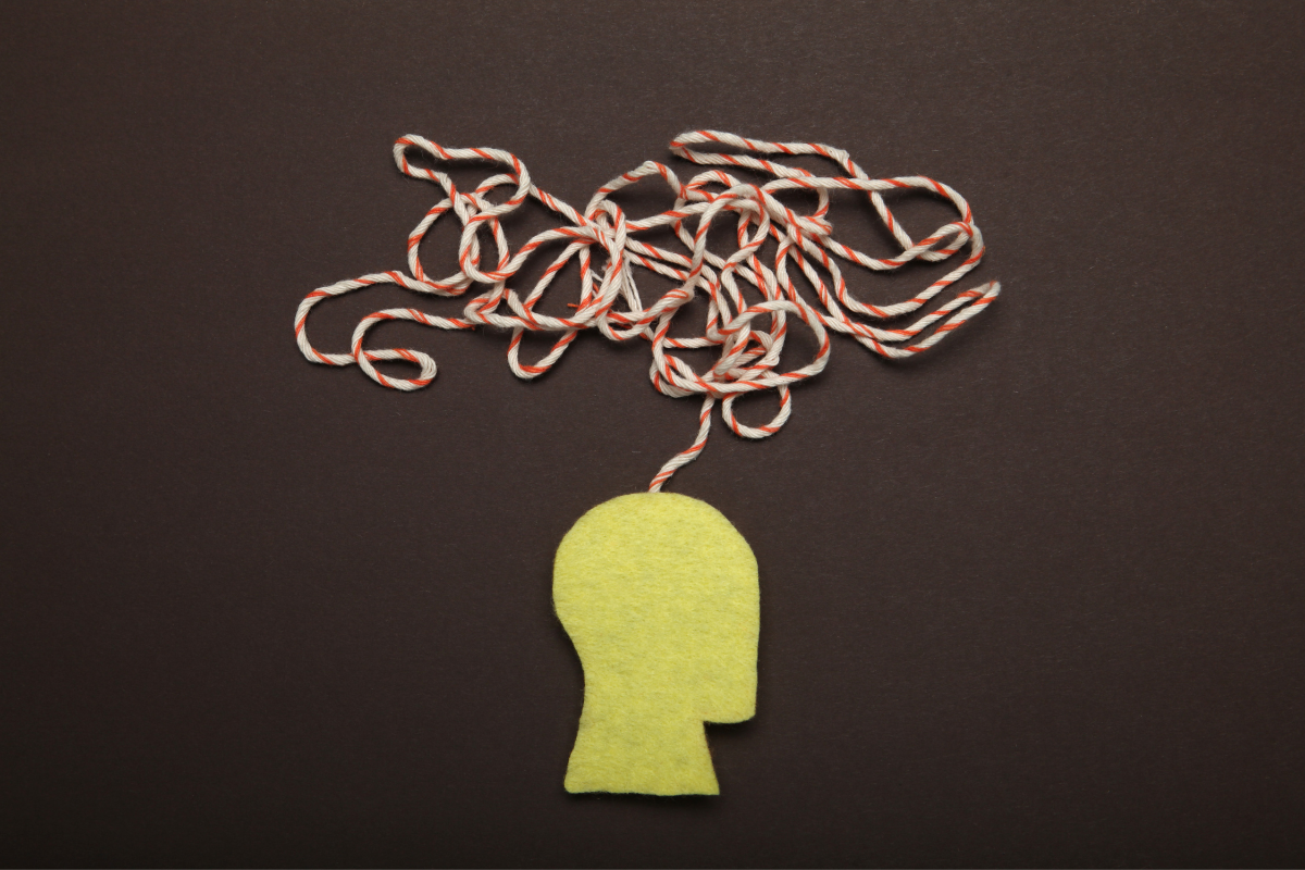 Une illustration symbolique montrant une silhouette de tête humaine en feutre jaune avec un fil rouge et blanc enchevêtré émergeant de son sommet, sur un fond marron foncé. Cela représente métaphoriquement la complexité des pensées et des émotions humaines durant la période de rentrée, et comment les compléments alimentaires, comme la créatine, peuvent être envisagés pour soutenir la santé mentale et cognitive des adultes face à ces défis.