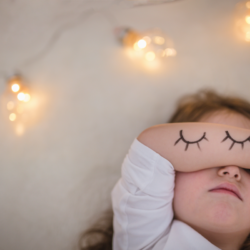 Une petite fille repose paisiblement, les yeux fermés, couverts par son bras sur lequel sont dessinés des cils, évoquant une scène de sommeil tranquille. L'ambiance est douce et relaxante, avec des lumières tamisées en arrière-plan qui suggèrent un environnement propice à un sommeil réparateur. Cette image symbolise l'importance du repos et le confort nécessaire pour un sommeil de qualité.