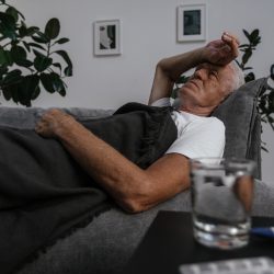 L'image représente un homme aux cheveux blancs sur son canapé. Il porte sa main à son front comme si il avait de la fièvre, symptôme des syndromes grippaux