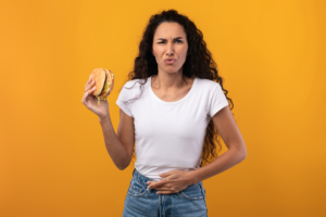 Une femme au visage contrarié tenant un hamburger dans une main, se tenant l’estomac de l’autre, exprimant un inconfort digestif, sur un fond jaune vif.
