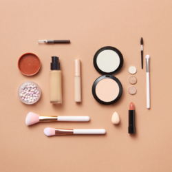 Ensemble de produits de maquillage disposés harmonieusement sur un fond uni beige. La sélection inclut un fond de teint liquide, un mascara, un correcteur en stick, une poudre compacte, un rouge à lèvres nude, des fards à paupières aux tons neutres, des perles de poudre multicolores, un blush en crème, ainsi que divers pinceaux de maquillage et une éponge à fond de teint.
