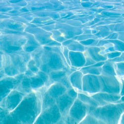 Vue en gros plan de la surface de l'eau d'un bleu clair et lumineux, avec des ondulations naturelles et des motifs ondulants créés par le mouvement doux des vagues sous un soleil éclatant, reflétant un spectre de couleurs comme des nuances de turquoise et des éclats de lumière arc-en-ciel.