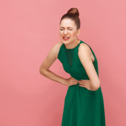 Femme souffrant de douleurs menstruelles avec les mains sur le ventre. Le fond rose contraste avec sa robe verte, mettant en évidence son expression de douleur. Ce visuel illustre l'article "Quelles sont les causes des règles douloureuses", abordant les causes courantes comme les contractions utérines intenses, l'endométriose, les fibromes utérins, et le syndrome prémenstruel (SPM).