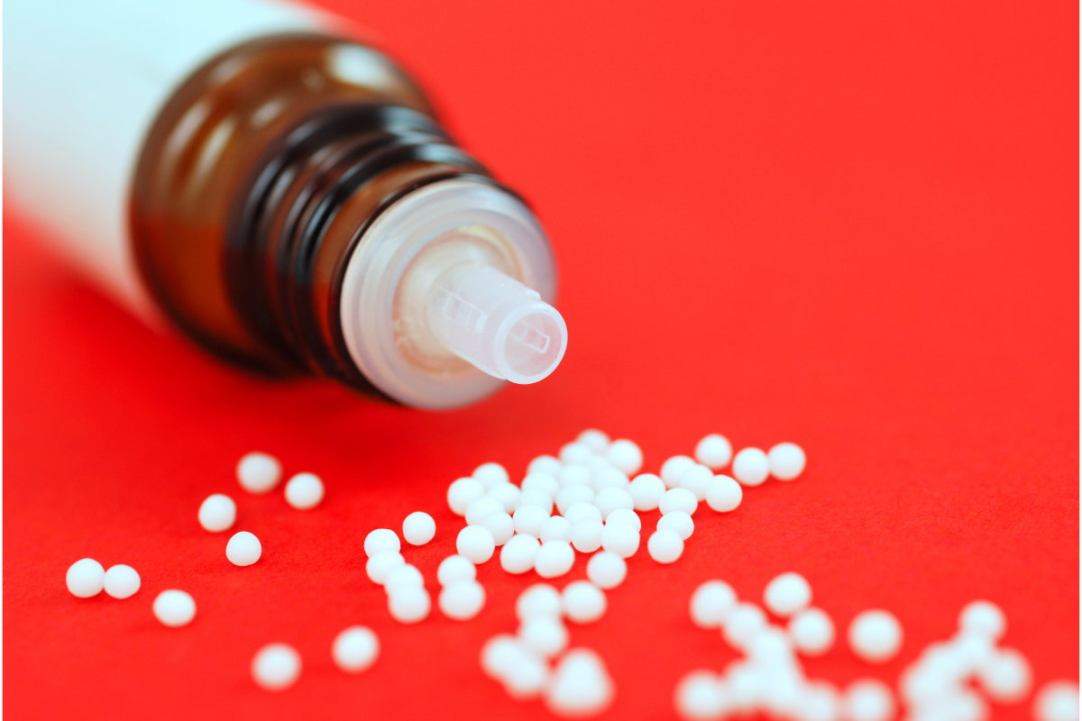 Flacon brun d'homéopathie avec capuchon blanc, renversé, laissant échapper des granules homéopathiques blancs sur un fond rouge. Ces granules sont utilisées pour soulager les traumatismes physiques tels que les bleus et les entorses, conformément aux pratiques homéopathiques traditionnelles.