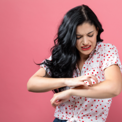 Sur cette photo, on voit une femme avec des cheveux longs et noirs, portant un haut à rayures horizontales noires et blanches. Elle regarde son bras droit avec une expression de douleur ou d'irritation, comme si elle avait une démangeaison ou une piqûre. Le fond est uni et rose, ce qui contraste avec son haut à rayures, mettant en évidence sa réaction. Son geste suggère qu'elle pourrait gratter ou frotter une zone irritée sur son poignet, ce qui est cohérent avec le sujet de l'eczéma ou d'autres affections cutanées irritantes.