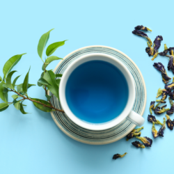 Tasse de tisane bleue avec des feuilles vertes et des pétales de fleurs séchées sur un fond bleu clair.