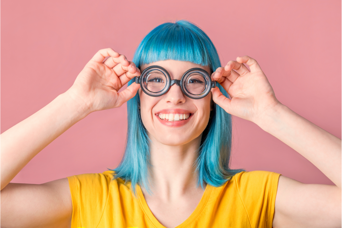 Femme souriante portant des lunettes épaisses et rondes, avec des cheveux bleus, sur fond rose.