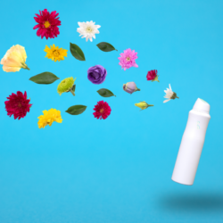Une bouteille de spray blanc avec le logo en vert est positionnée à droite sur un fond bleu clair. À gauche, un assortiment de fleurs colorées en lévitation, incluant des dahlias rouges, des chrysanthèmes jaunes, des roses, et des fleurs de passiflore, symbolisant les ingrédients naturels utilisés dans le spray.