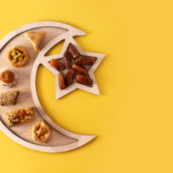 Cette image présente un assortiment de pâtisseries et de dattes disposées artistiquement sur une planche à découper en bois dont la forme évoque le croissant de lune et l'étoile, symboles souvent associés à l'Islam. Le fond jaune vif contraste avec les couleurs naturelles des aliments, mettant en valeur les textures et les formes variées des douceurs traditionnellement consommées pendant le Ramadan. Cela pourrait accompagner un article traitant des traditions culinaires du Ramadan 2024, en mettant l'accent sur la rupture du jeûne et les moments de partage qui caractérisent ce mois sacré.
