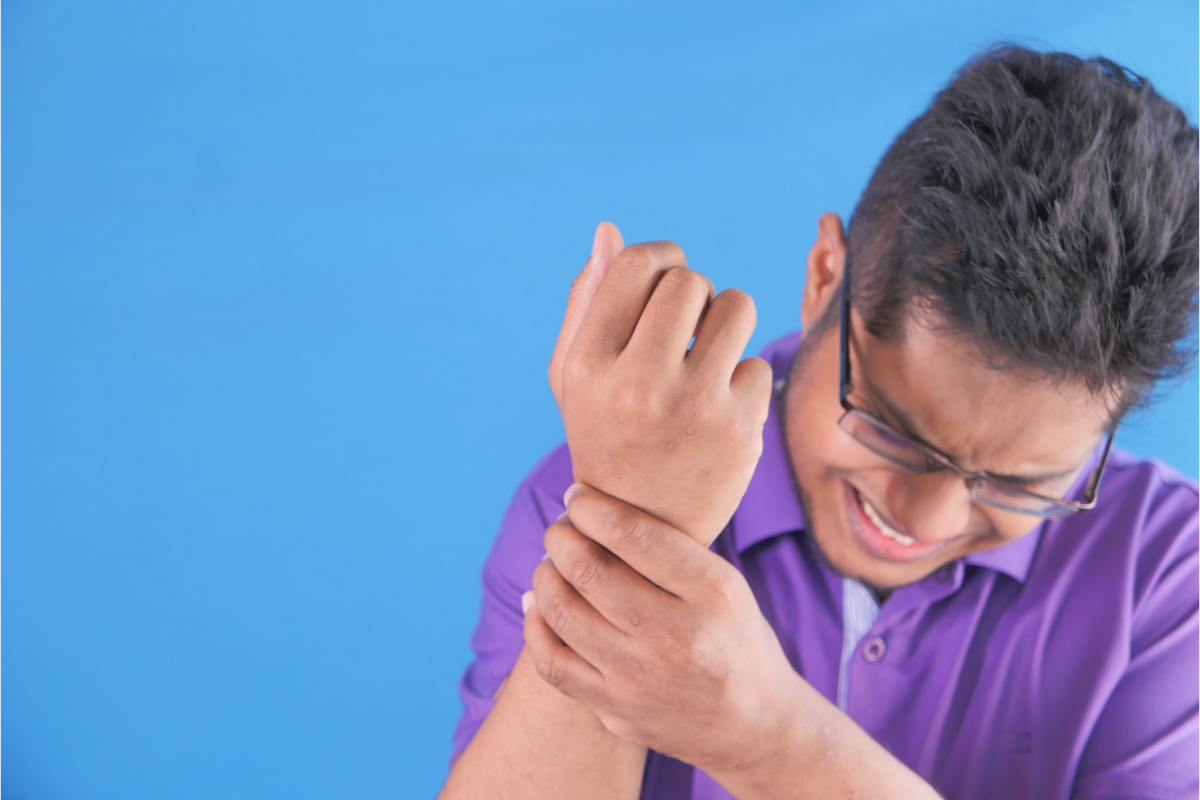 Un homme semble éprouver une vive douleur au poignet gauche, qu'il tient fermement avec sa main droite. Il porte des lunettes et une chemise mauve, et grimace de douleur. Le fond de l'image est uni et bleu, mettant en évidence la réaction de l'homme à la douleur. Cela pourrait illustrer un article sur la gestion de la douleur, notamment par des méthodes comme l'homéopathie.