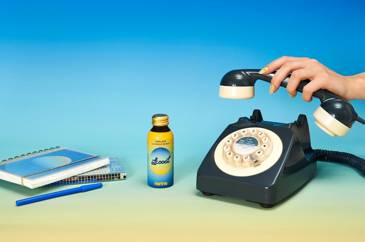 L'image montre une composition aux couleurs vives avec un fond dégradé allant du bleu clair au jaune. Au premier plan, à gauche, il y a un carnet à spirales ouvert sur une page vierge avec un stylo bleu posé dessus. À côté, un petit flacon d'alcool de couleur jaune avec l'inscription "alcool nonna" et une étiquette qui suggère une utilisation pour "soulager la gueule de bois". Sur la droite, un téléphone à cadran rotatif de couleur noire, avec le combiné soulevé par une main féminine décorée d'ongles vernis aux motifs colorés. Le style évoque un bureau vintage ou une scène d'un film rétro. Nonna Lab : Une révolution dans le monde des compléments alimentaires