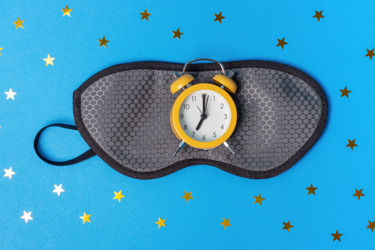 Un masque de sommeil noir avec un réveil jaune posé dessus, sur fond bleu orné d'étoiles dorées.