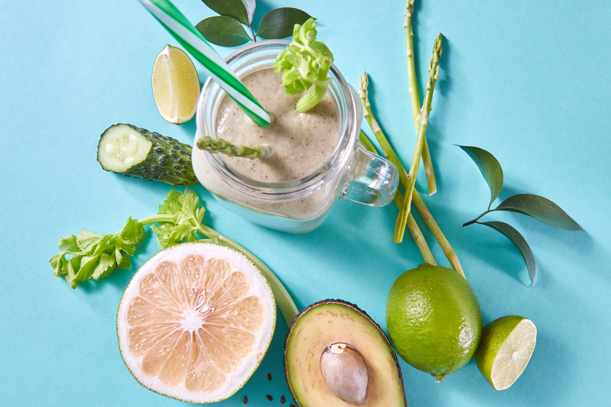 Une composition saine et détoxifiante centrée sur un smoothie vert crémeux dans un bocal en verre avec une paille, entouré d'ingrédients naturels et frais. On y trouve des tranches de concombre, une demi-avocat avec son noyau, un citron vert entier et coupé en deux, ainsi que des asperges fraîches et des feuilles de céleri. Le tout est disposé harmonieusement sur un fond bleu turquoise, évoquant une atmosphère pure et vivifiante, parfaitement en ligne avec le thème de la détoxification par l'homéopathie.