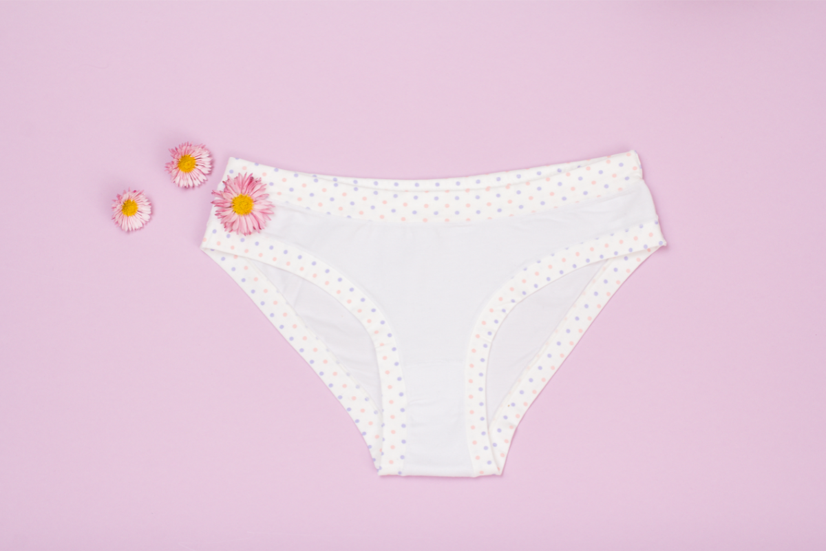 Une culotte blanche avec des pois colorés, accompagnée de petites fleurs roses, sur un fond rose clair. Cette image illustre le sujet de la leucorrhée, mettant en avant l'importance de l'hygiène intime et les remèdes naturels comme l'homéopathie pour traiter ce trouble.
