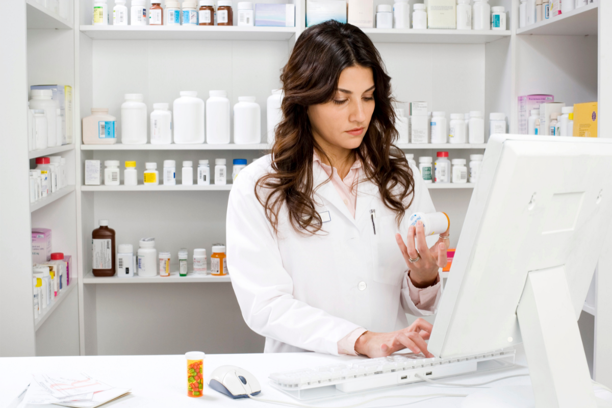 Une pharmacienne examine un médicament tout en travaillant sur un ordinateur dans une pharmacie. Cette image illustre le rôle crucial du pharmacien dans l'accompagnement des patients en surpoids ou obèses, notamment par des conseils nutritionnels et la recommandation de compléments alimentaires pour améliorer la santé intestinale et gérer le poids.
