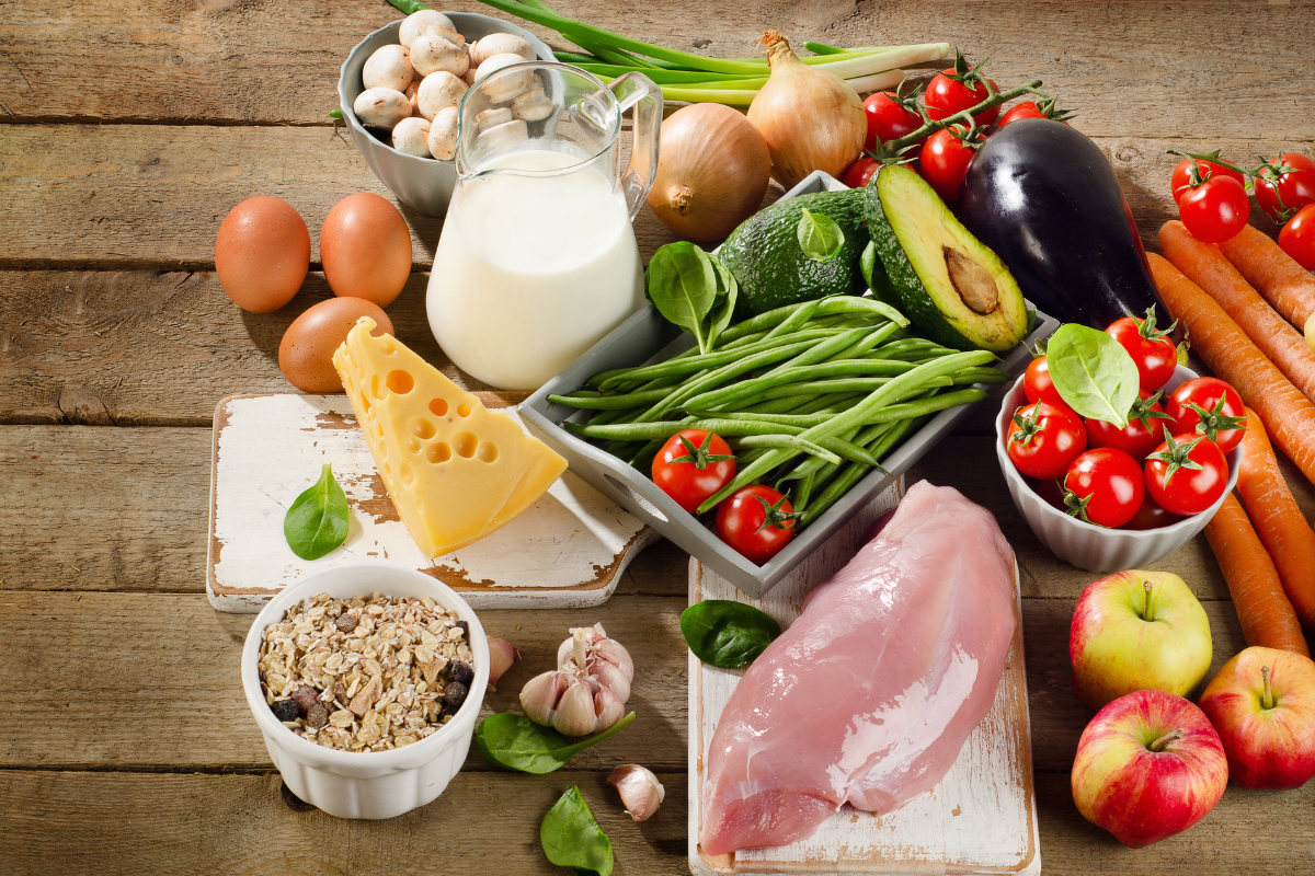 Aliments frais et sains représentant différents régimes alimentaires : légumes verts, tomates cerises, avocat, œufs, fromage, lait, poulet, céréales et fruits, illustrant l’influence de la nutrition sur le microbiote intestinal.