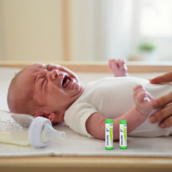 Bébé allongé et pleurant, avec un biberon et deux tubes d’homéopathie à proximité, illustrant les solutions naturelles contre les coliques du nourrisson.