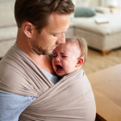 Un père porte et berce un bébé qui pleure pour le soulager d’une crise de coliques, avec un biberon posé sur la table à proximité.
